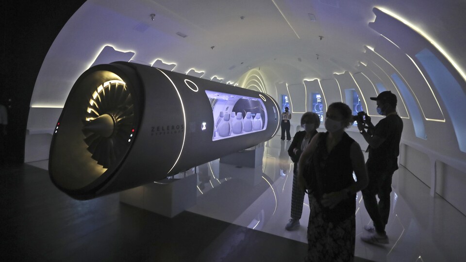 Trois personnes en silhouette observent un véhicule Hyperloop dans une salle d'exposition.