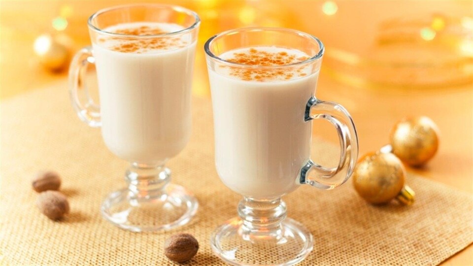 Deux tasses de lait de poule sont sur une table décorée pour Noël avec des noisettes, deux boules dorées et un napperon doré.