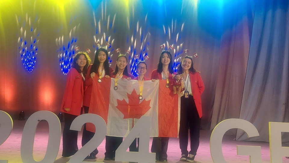 Six jeunes femmes avec des médailles et un toutou d'orignal tiennent un drapeau canadien et portent des serre-têtes avec des drapeaux canadiens.