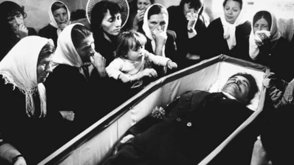 Une photo en noir et blanc d'un homme dans un cerceuil entouré de personnes en deuil.