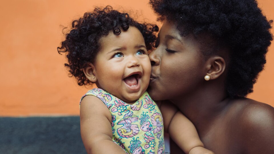 La fillette a la peau métisse et des frisettes, la maman a la peau noire et une coupe afro courte. La maman fait un bisou à son enfant qui sourit. 
