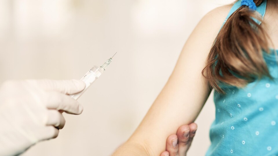 Une main gantée approche une seringue du bras d'une petite fille habillée en bleu.