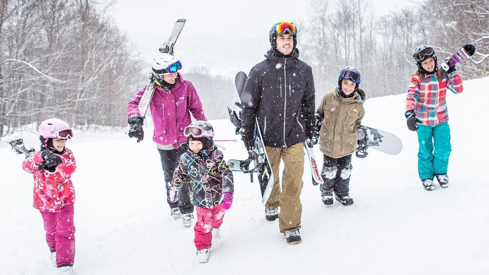 Deux adultes et quatre enfants marchent dans la neige en portant leurs skis et leurs planches à neige.