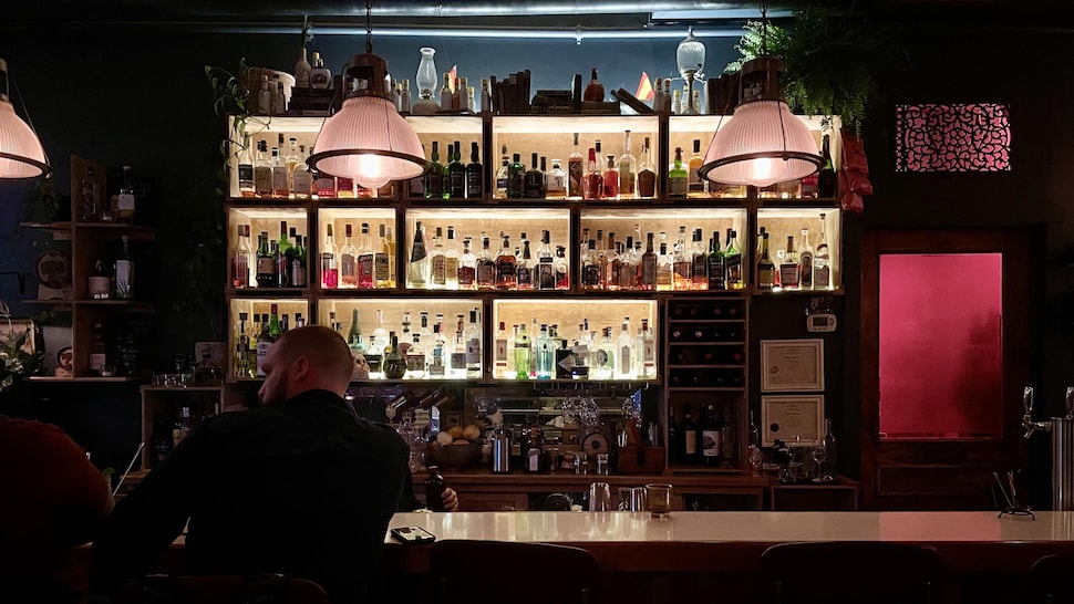 Un bar sombre avec des rangées de bouteilles, sous un lumière tamisée rosée. 