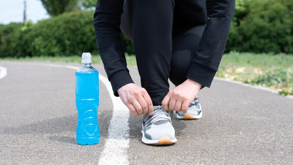 Une bouteille remplie d'une boisson sportive bleue posée au sol à côté du pied d'une personne qui noue le lacet d'un soulier, en pause durant une course.
