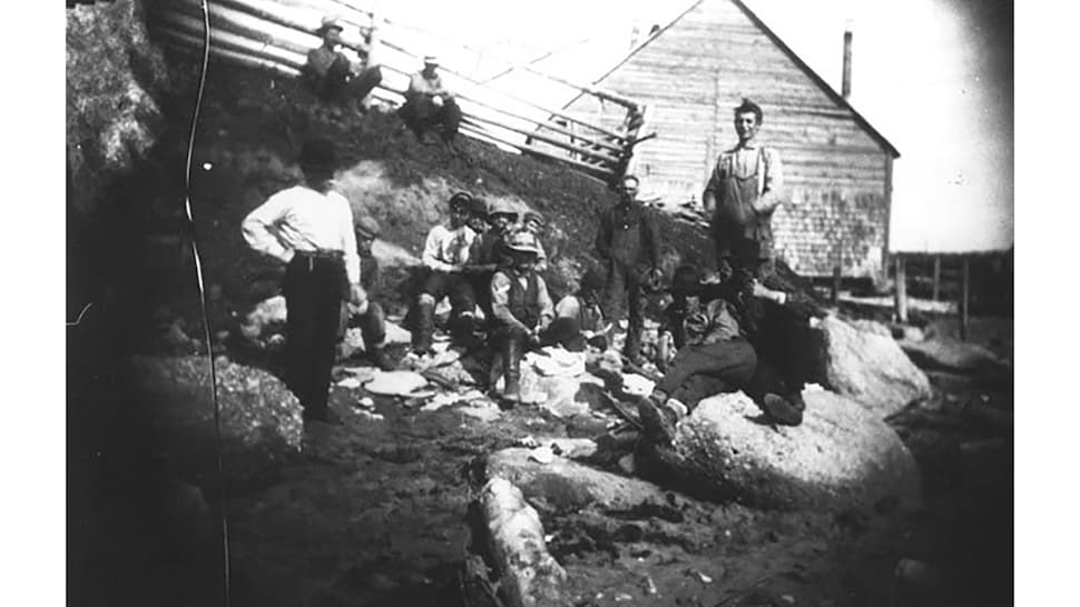 Une photo en noir et blanc avec des employés de la conserverie qui prennent une pause devant l'entreprise en bois.