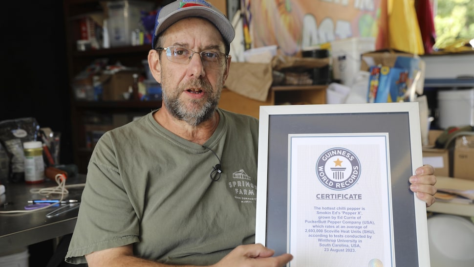 Un homme portant un t-shirt et une casquette est assis dans un atelier et tient dans ses mains un certificat encadré.