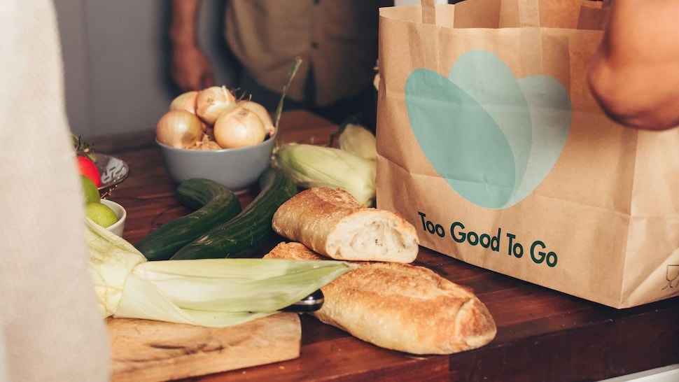Un sac avec le logo de la marque est déposé sur une table de bois avec plusieurs aliments, comme du pain et des légumes.