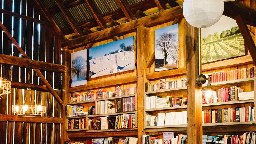 Sur cette photo, il est possible de voir un mur seulement de la grange, où sont entassé plusieurs livres sur différents étages . Au pied du mur, il y a trois tables où l'on peut s'installer pour lire. 
