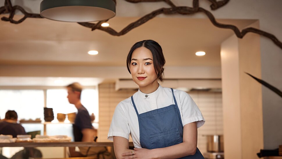 Photo de la femme dans un restaurant le jour. Elle porte un tablier bleu et se tient les bras croisés.