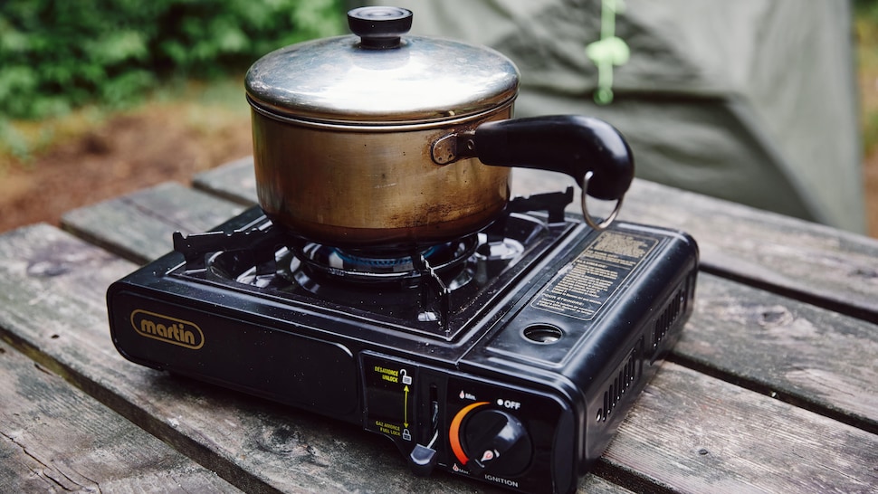 Sur une table en bois, est déposé une cuisinière portative.  Une petite casserole en métal, est déposé sur la cuisinière portative.