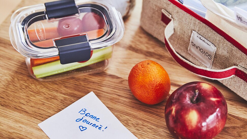 Un contenant avec des crudités, une clémentine, une pomme et une boîte à lunch ouverte sont sur une table, accompagnés d’un mot sur lequel est écrit bonne journée. 