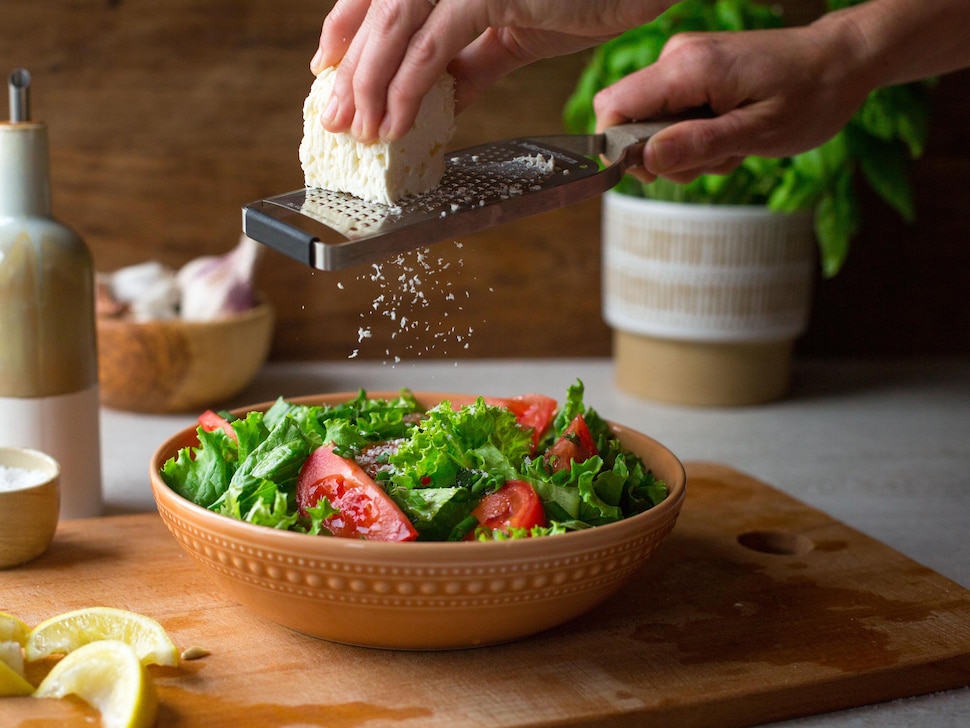 Une personne râpe du fromage feta sur une salade. La portion individuelle est servie dans un bol de terre cuite. 