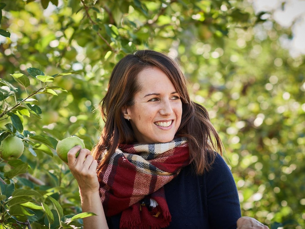 Geneviève O'Gleman se trouve dans un verger et tient une pomme croquée. 