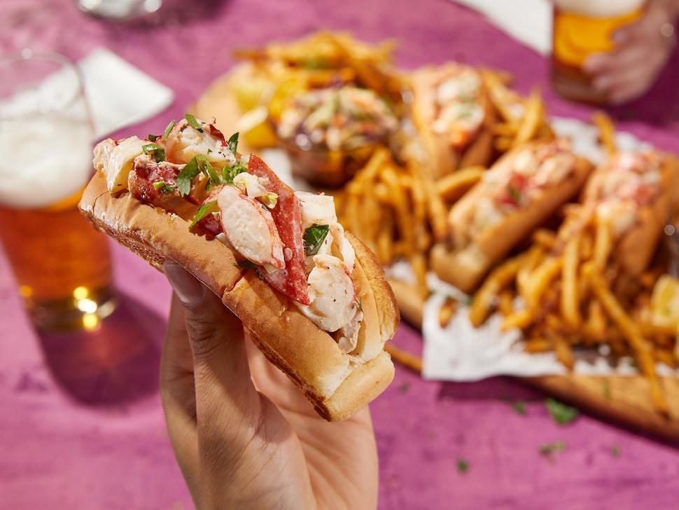 Une main tient un pain hot-dog farci de homard, en arrière-plan, un plateau de frites et de guédilles de homard. 