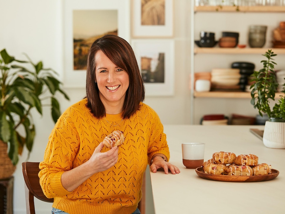 La nutritionniste Geneviève O’Gleman est assise près du comptoir de la cuisine et tient un beignet aux pommes. Sur le comptoir, il y a une assiette de beignets aux pommes, une tasse de thé et des plantes. 