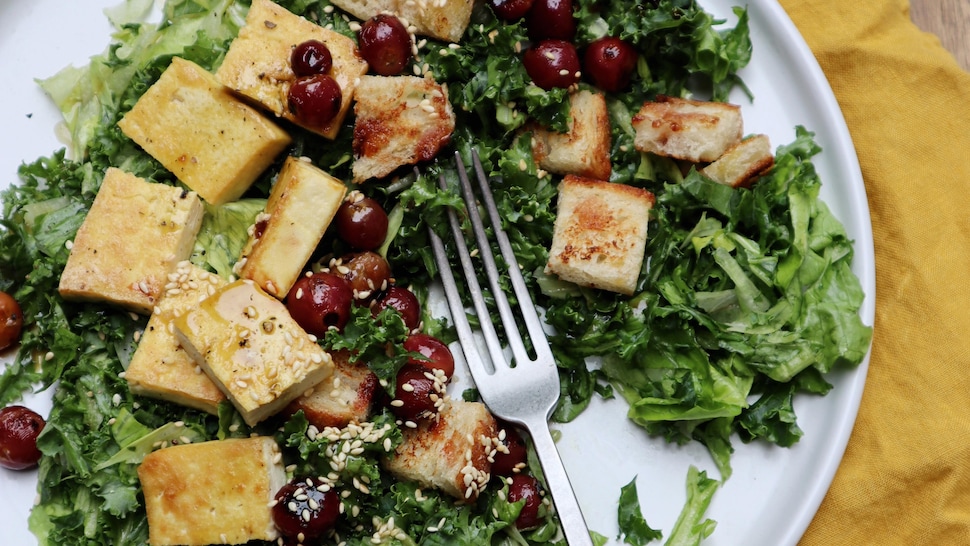 Dans une assiette, il y a une salade verte garnie de morceaux de tofu grillés, des raisins et des croûtons.