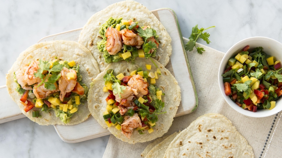 Une planche de bois sur laquelle il y a trois tacos aux crevettes, à la salsa et à la mangue.