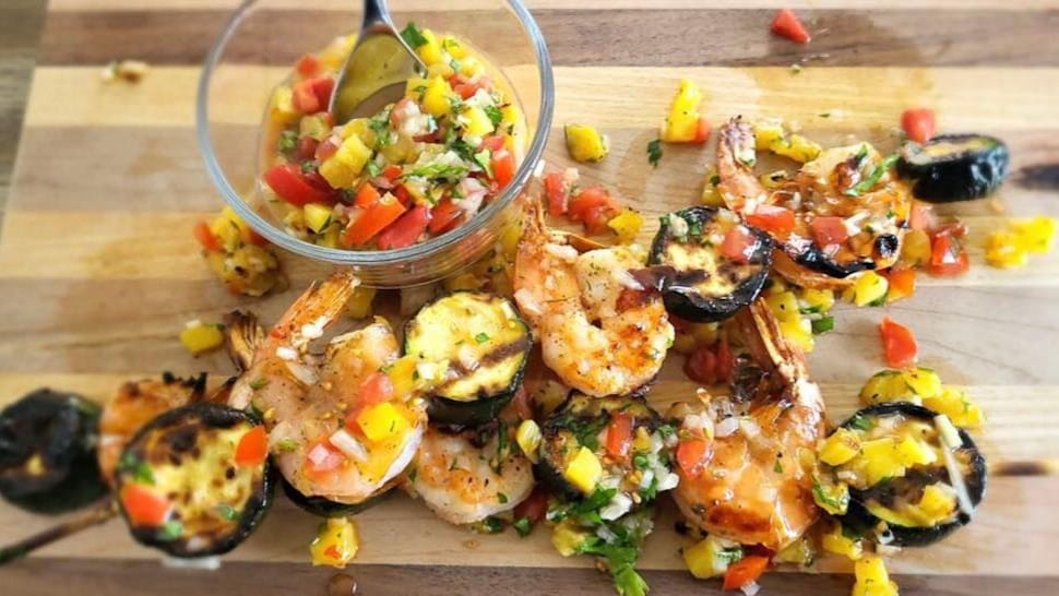 Deux brochettes de crevettes et de courgettes recouvertes de salsa à l'ananas sur une planche.