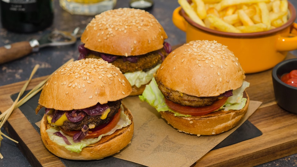 Trois burgers sur une planche en bois avec un bol de frites en arrière-plan.