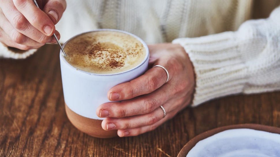 Une main tient un gobelet bleu pâle contenant un café latté à la citrouille et l'autre main tiens une cuillère pour brasser.