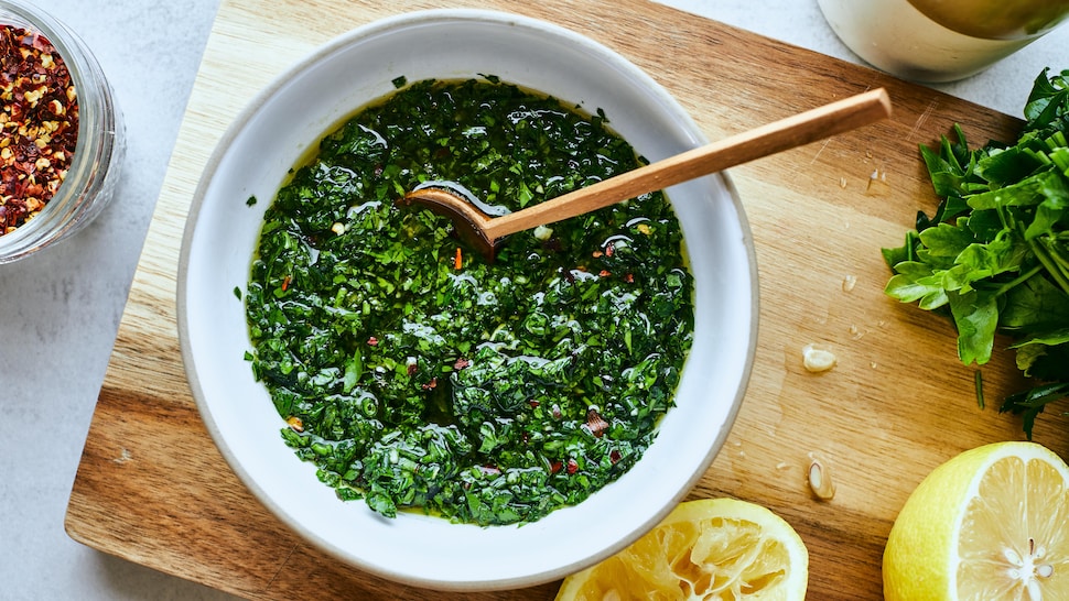 Un bol rempli de chimichurri sur une planche de bois. Des citrons, du persil, de l'huile d'olive et des flocons de piments sont placés autour. 