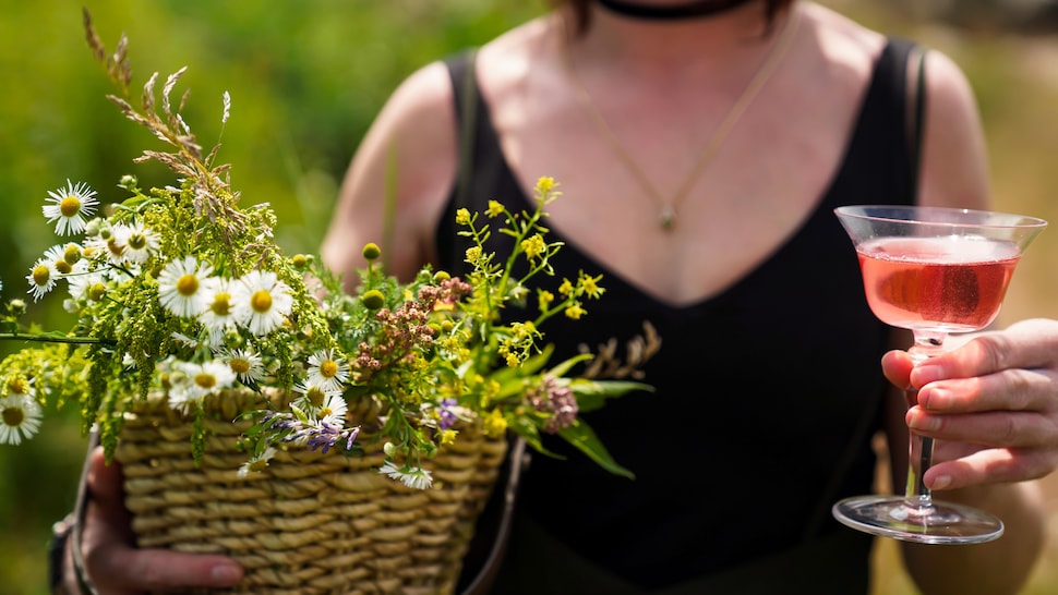 Une femme tenant un panier de fleurs sauvages dans une main et un cocktail dans l'autre. 