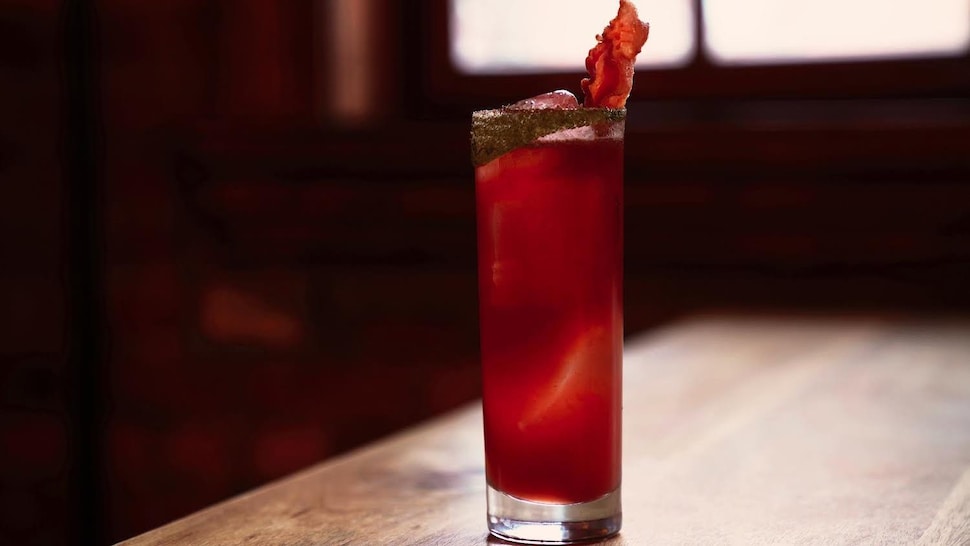 Un cocktail rouge dans un verre allongé avec une tranche de bacon en décoration.