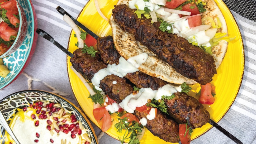 Des kebabs dans une assiette avec un pain pita, des morceaux de tomates et d'oignons et de la sauce.