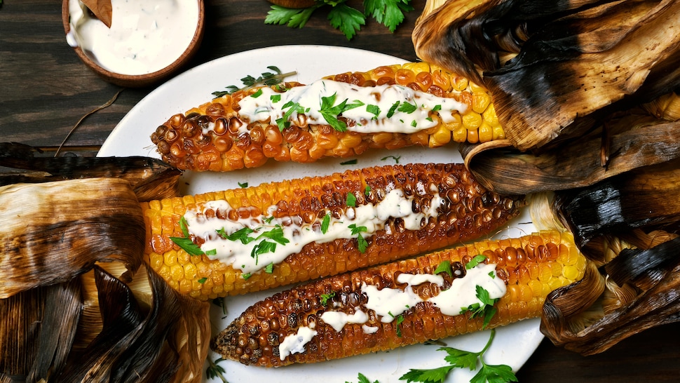 Des épis de maïs grillés avec leur feuille, de la mayo sur une assiette.