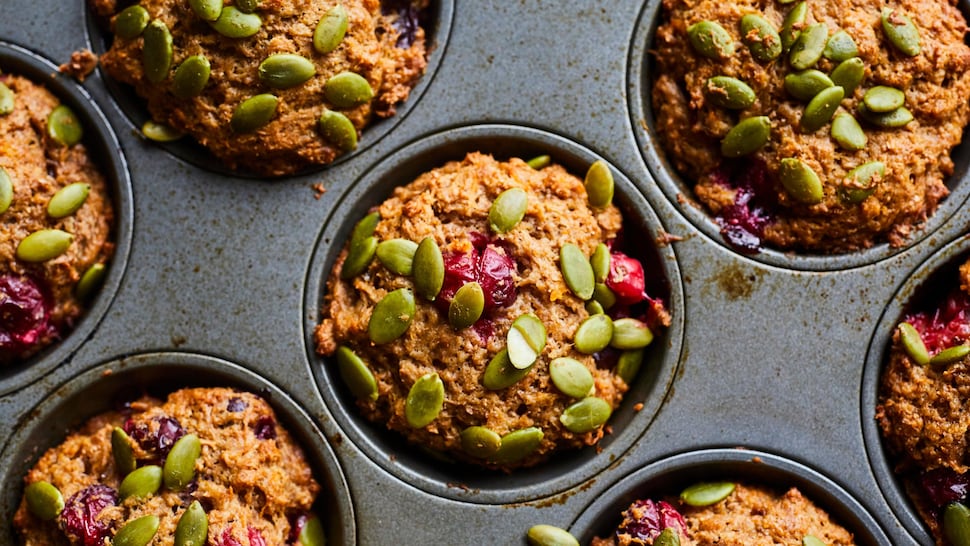 Plusieurs muffins à la patate douce et aux canneberges dans une plaque à muffins, recouvert de graines de citrouille.