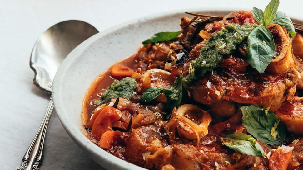 Un bol d'osso buco nappé de sauce, garni de fines herbes et servi avec une cuillère en argenterie.