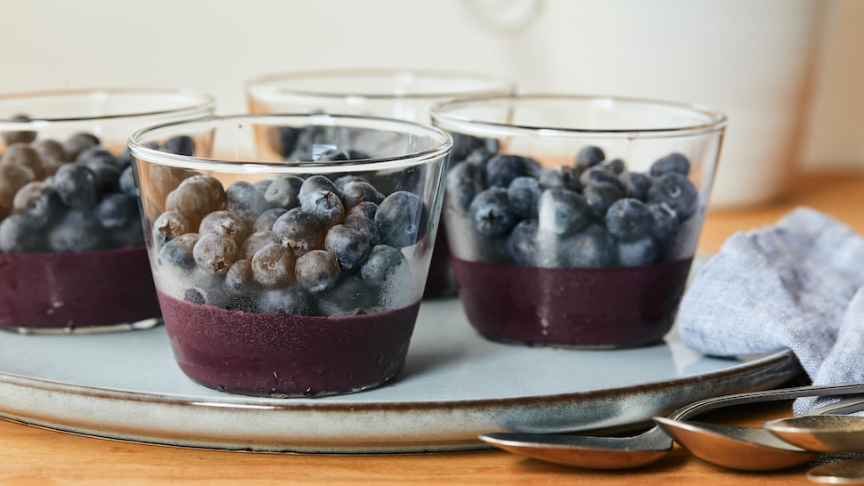 Sur une assiette, est déposé quatre petits pots en verre, contenant la recette de choco-bleuets glacés au fond et des bleuets frais sur le dessus.