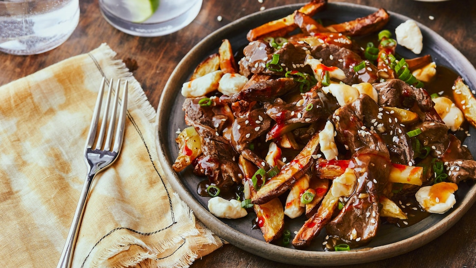 Une poutine recouverte de bulgogi de bœuf est dans une assiette, accompagnée d’une sauce au gochujang, d’oignons verts et de graines de sésame.