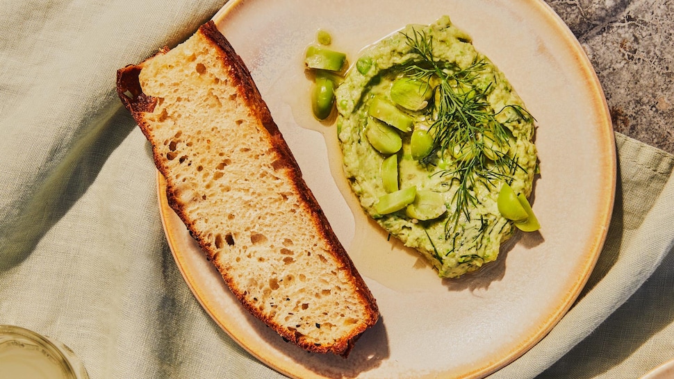 Dans une assiette, il y a de la purée de gourganes fraîches avec un croûton de pain.