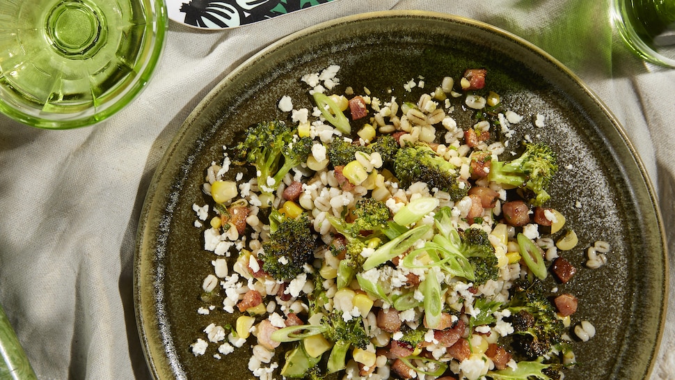 Un plat de salade de brocoli, bacon, maïs, orge et feta sur une table.