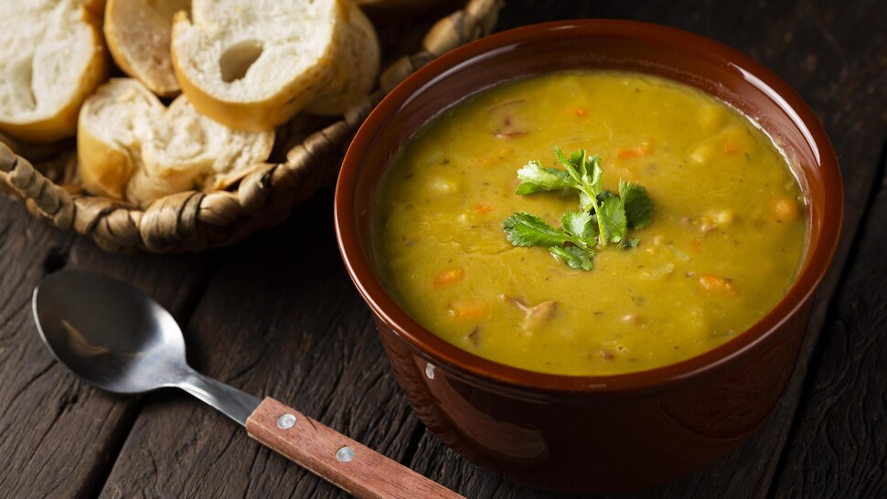 Un bol de soupe aux pois, un panier rempli de pain baquette en tranches et une cuillère sur une table en bois.