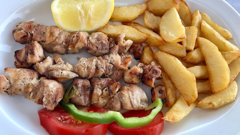 Des souvlakis au poulet dans une assiette avec des frites, des morceaux de tomates, de poivrons et du citron.