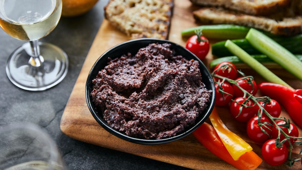 Bol de tapenade aux olives Kalamata sur une planche de bois avec des légumes, du pain grillé et une coupe de vin blanc.