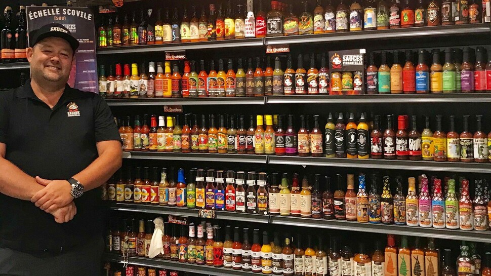 Un homme sourit à la caméra à côté d'un étalage présentant des dizaines de bouteilles de sauces piquantes.
