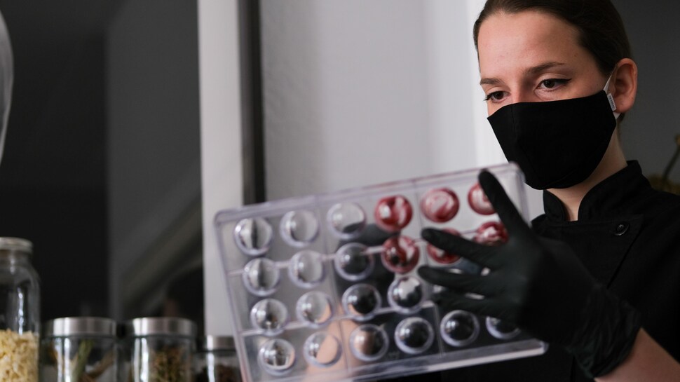 Une femme porte un masque au visage et peint un moule de chocolat avec son doigt ganté. 