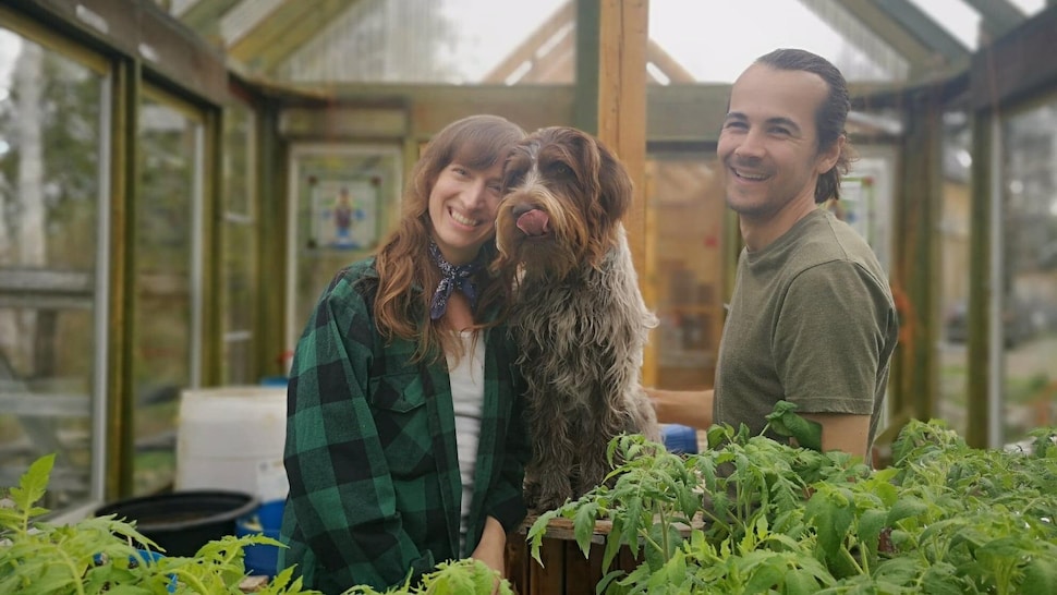 Dans une serre, Eve-Andrée et Philippe regardent vers l'objectif en souriant, alors que Thelma le chien se lèche les babines.