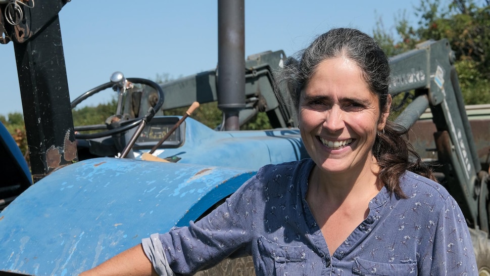 Une femme devant son tracteur bleu, un peu éblouie par le soleil, sourit à la caméra.