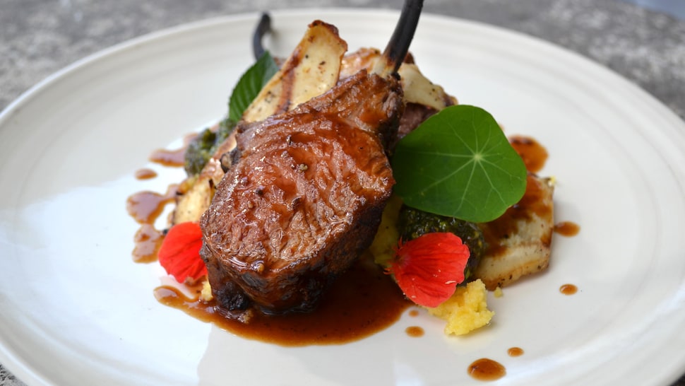 Carré d’agneau au barbecue, tapenade de pistaches et parmesan présenté dans une assiette blanche.