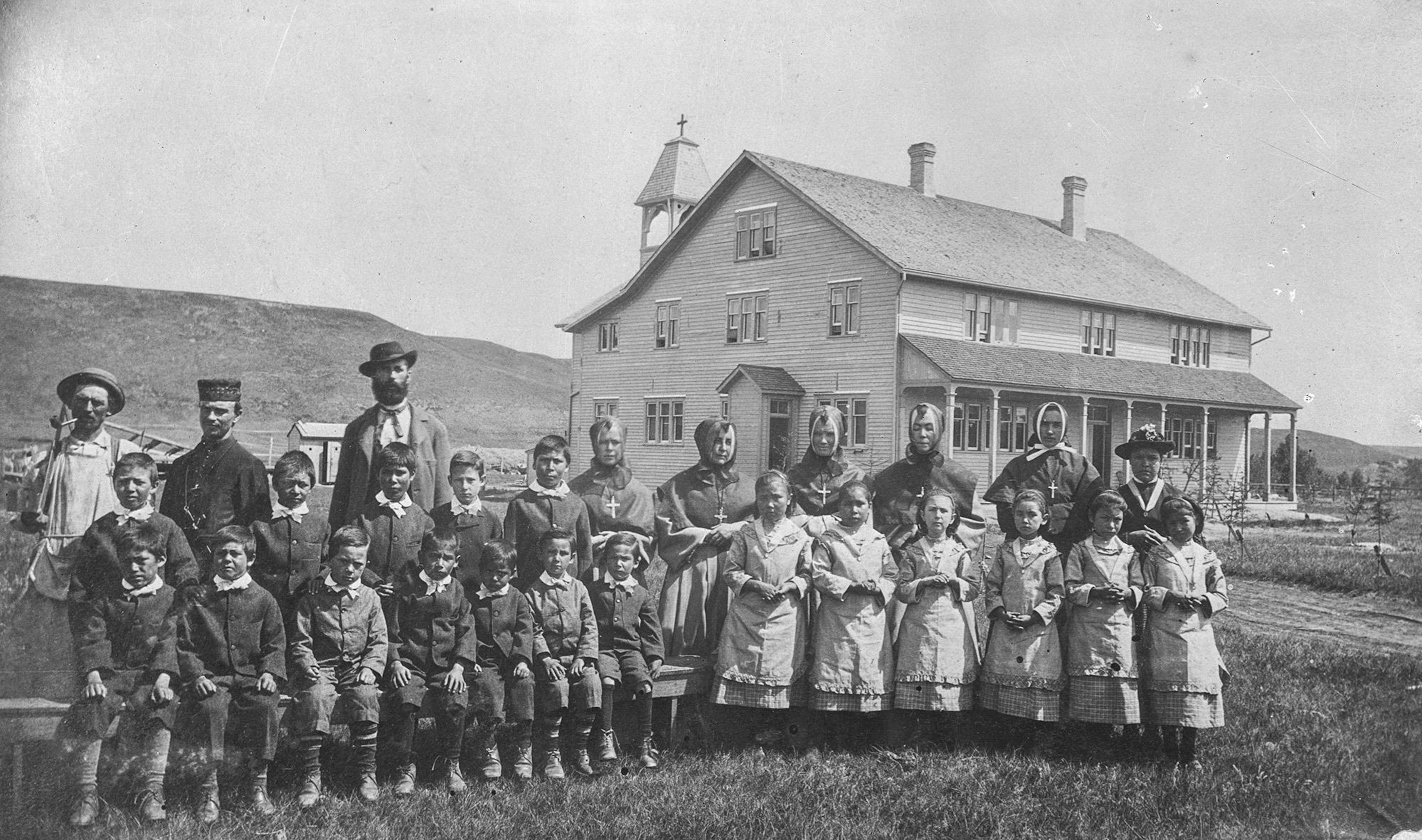 Photo d'archive où l'on voit des élèves et leurs superviseurs regroupés sur le terrain devant l'édifice. Des élèves garçons à gauche, des élèves filles à droite.