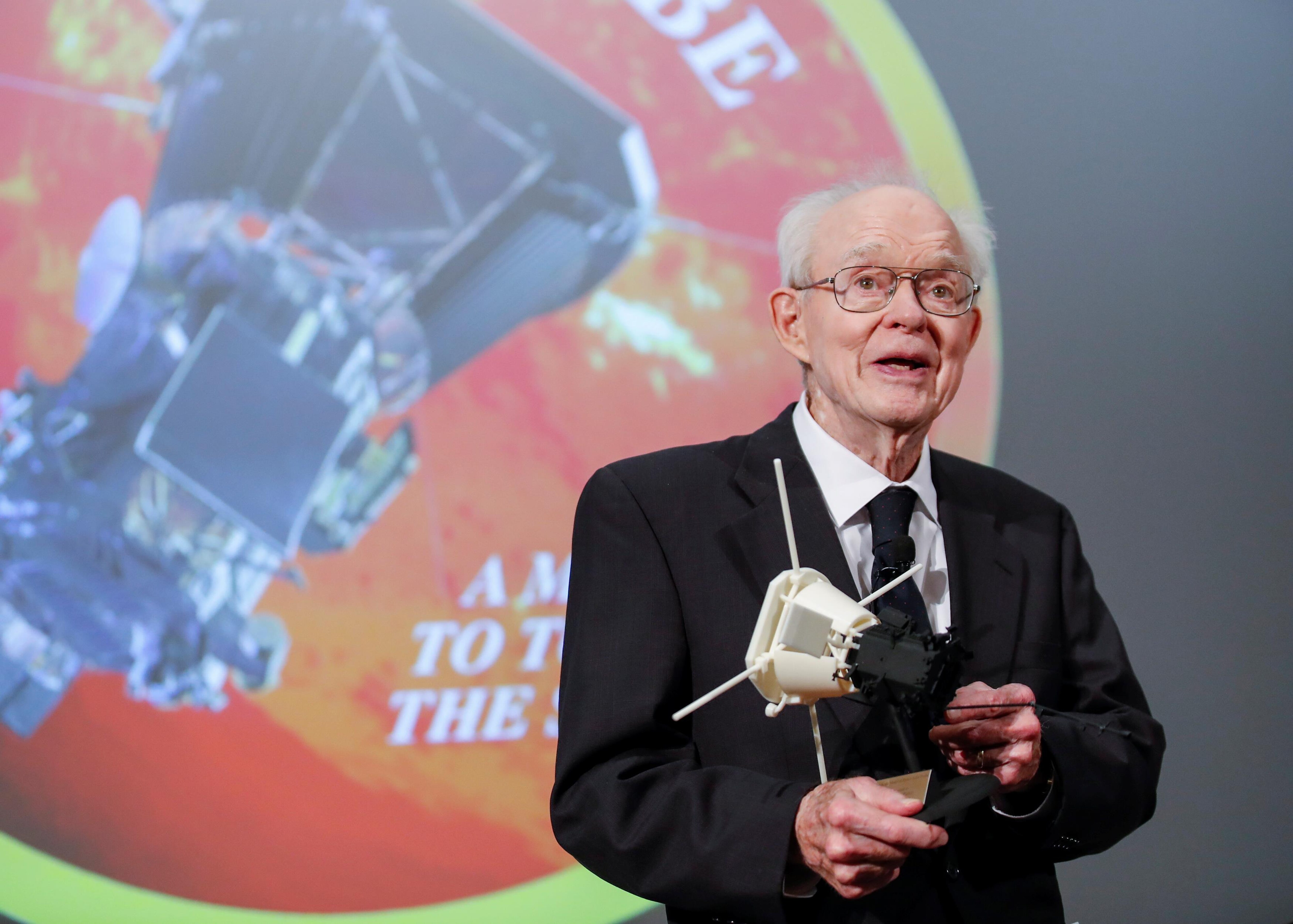 Un scientifique tenant une maquette devant une projection d'un logo de mission spatiale.