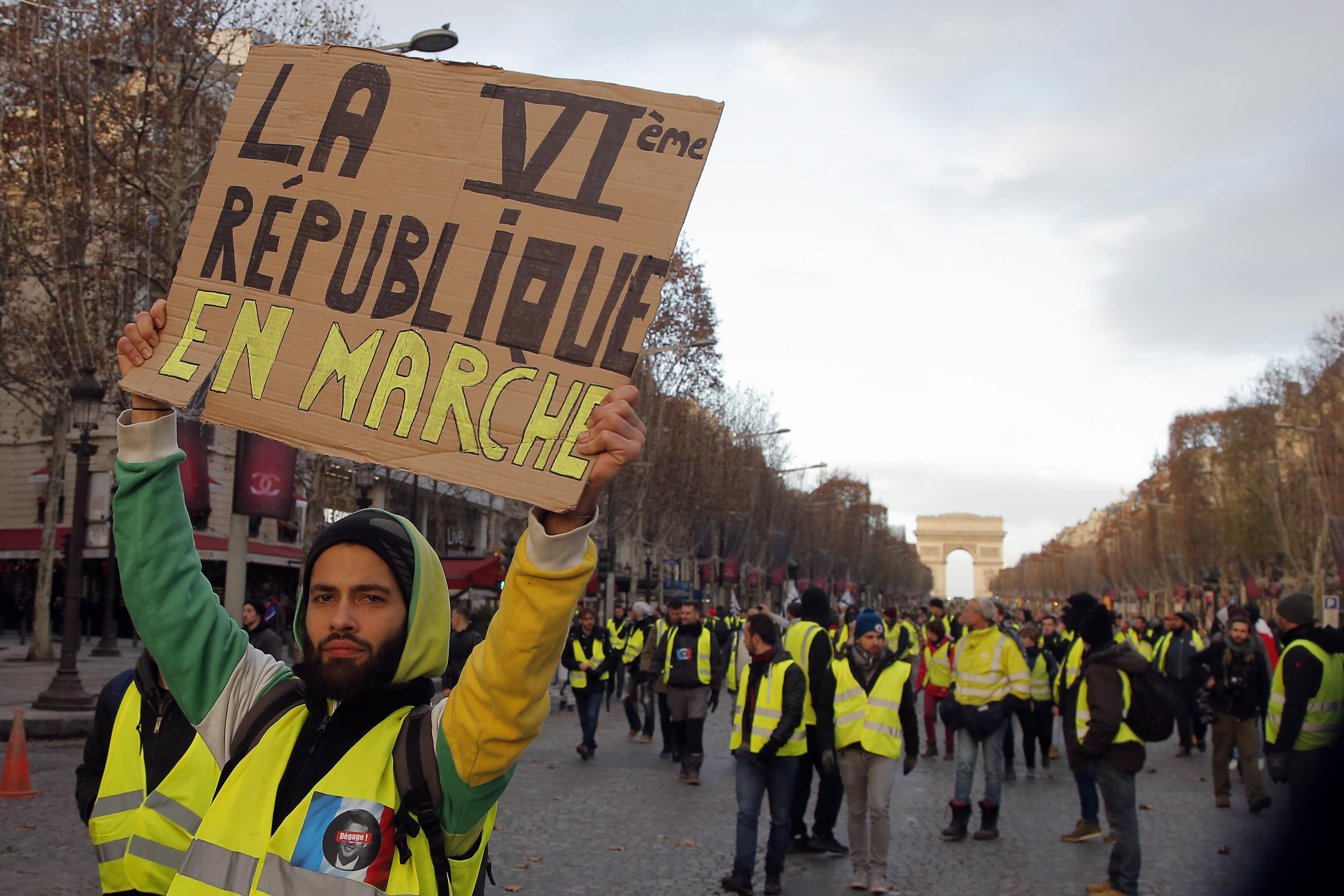 Sur les Champs-Élysées, à Paris, un manifestant portant un gilet jaune tient une pancarte en carton où il est écrit « La sixième République en marche ».