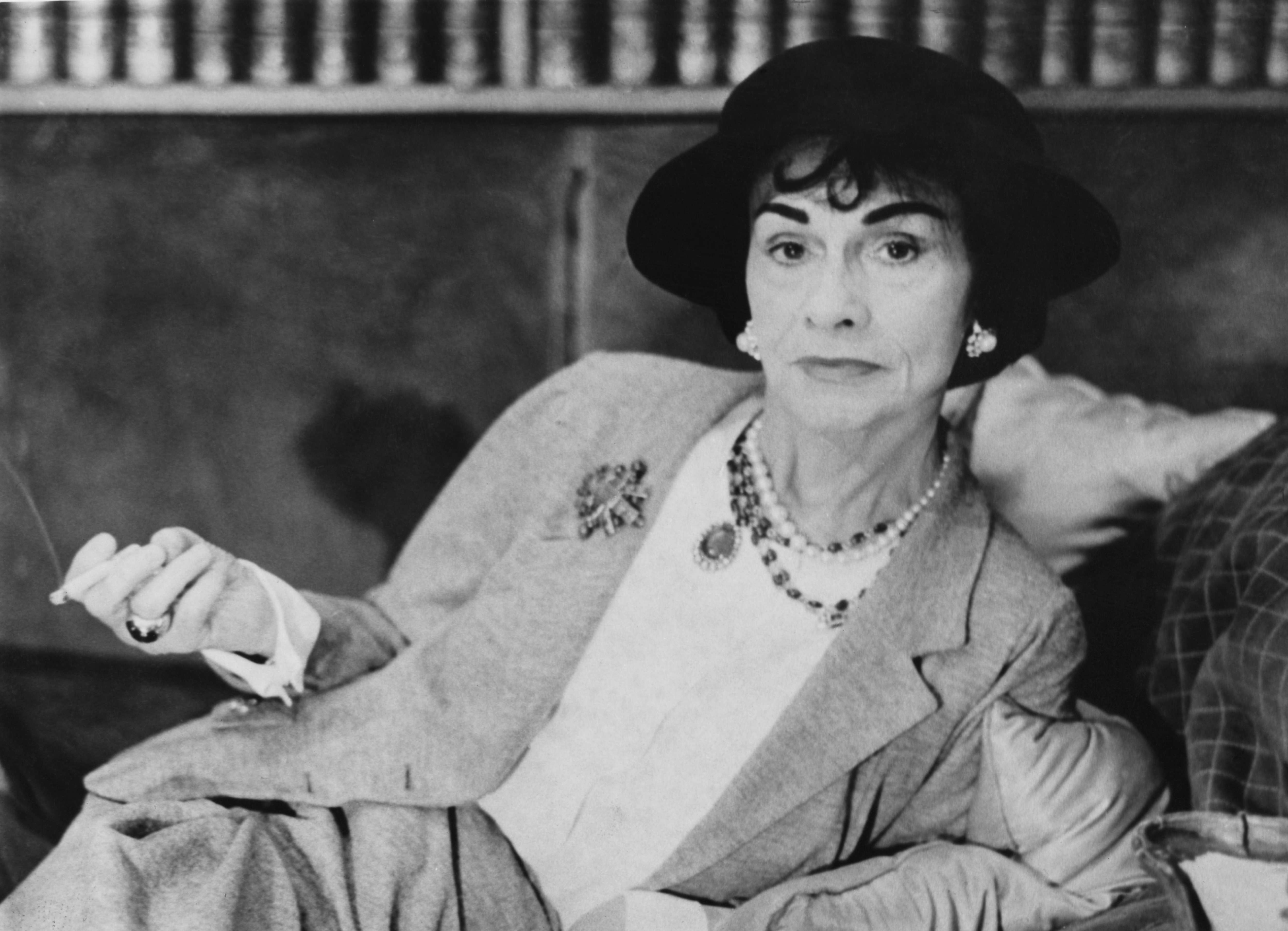 Coco Chanel porte des bijoux, un tailleur et un chapeau.