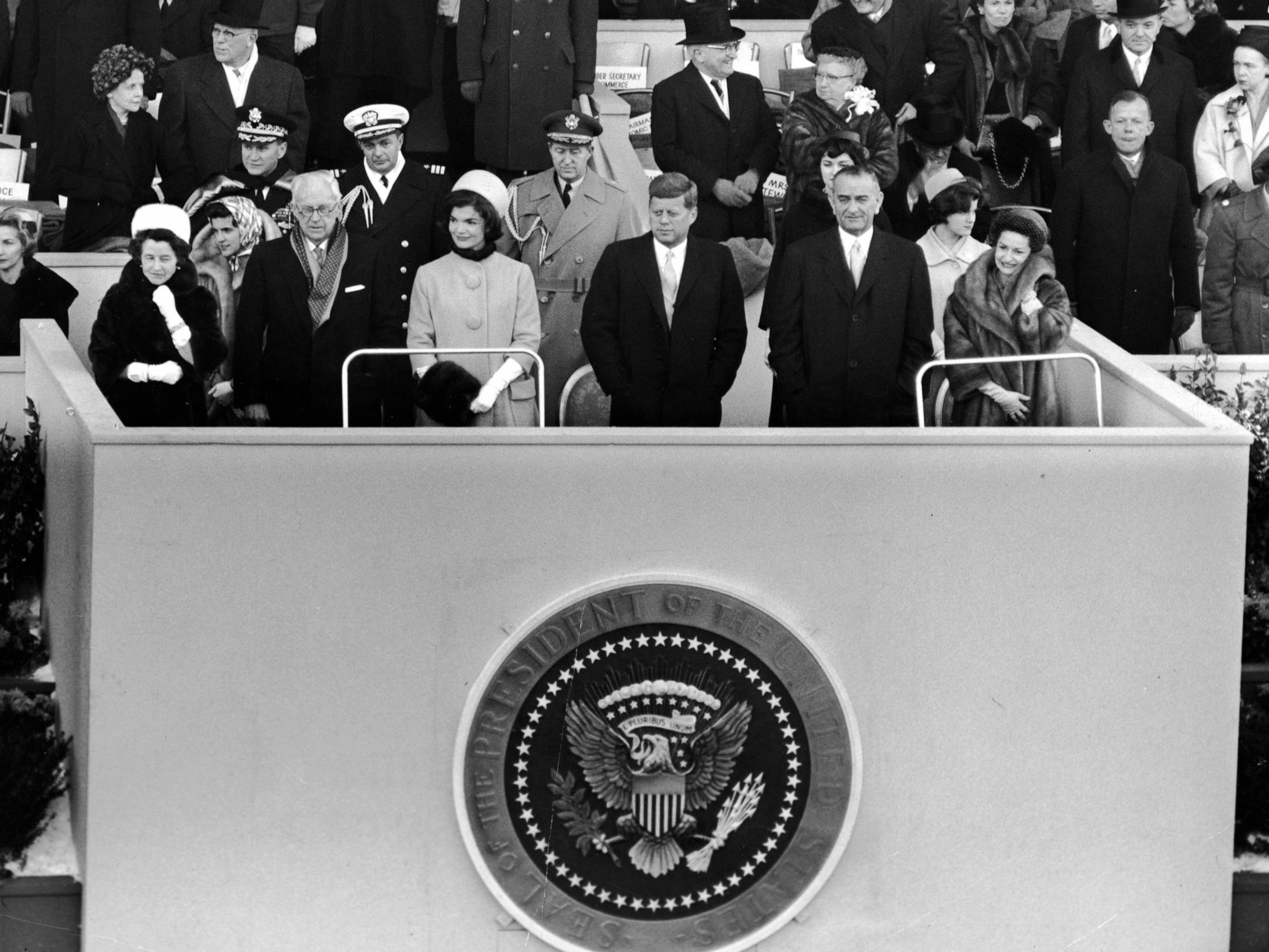 Photo en noir et blanc d'une douzaine de personnes debout sur une plateforme qui porte le sigle du président des États-Unis.