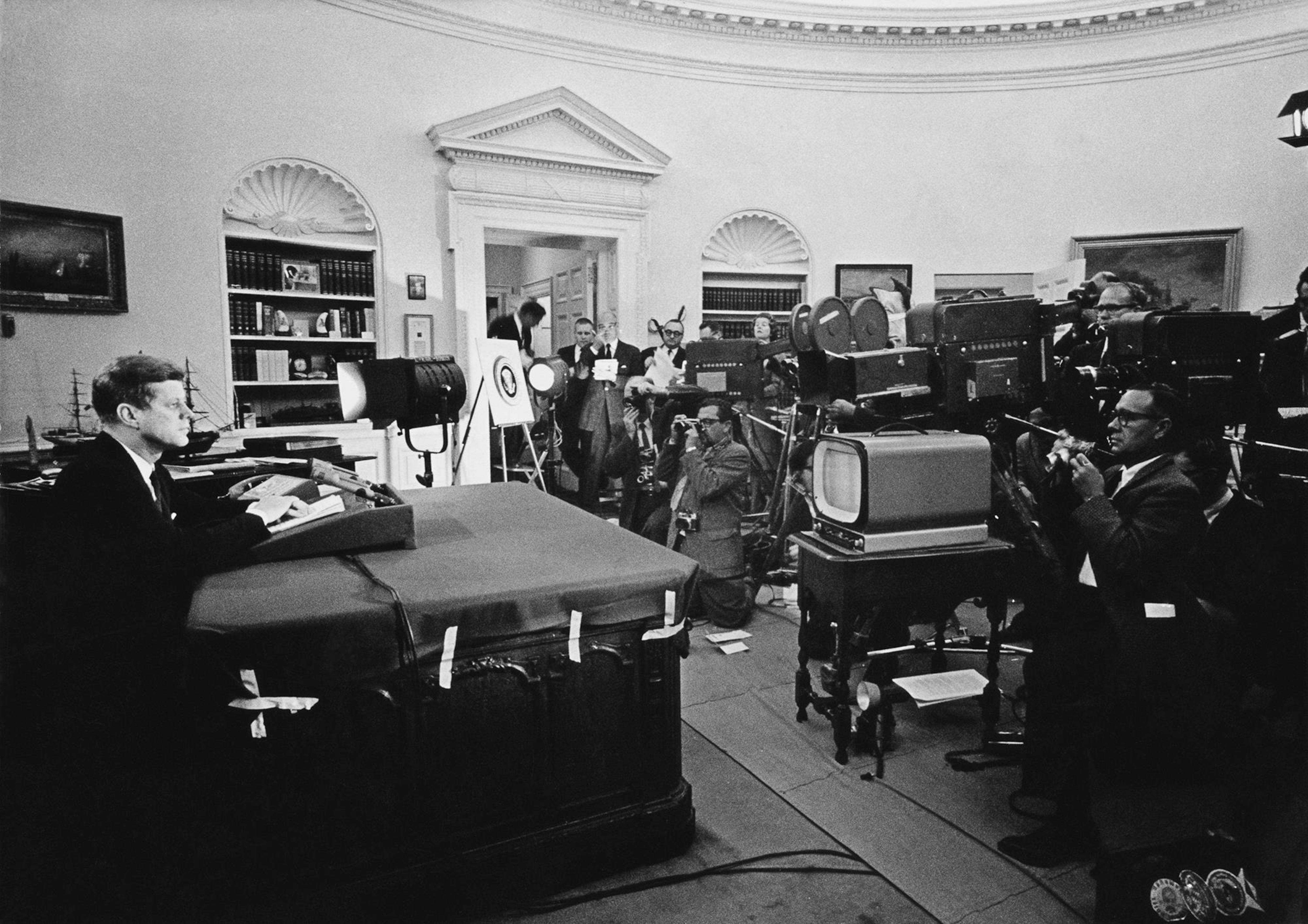 Photo en noir et blanc d'un homme assis devant un bureau. Une nuée de caméras de télévision et quelques photographes sont devant lui.